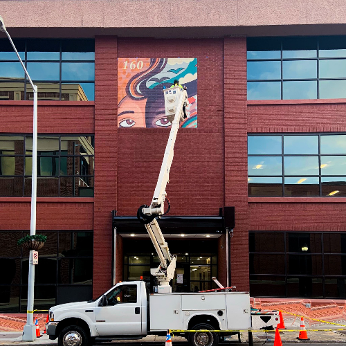 A photo of a truck with an electric lift applying a graphic on the side of a building.