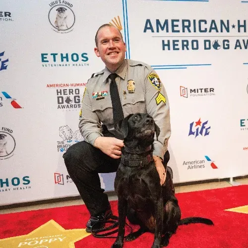 Man in uniform poses next to dog on red carpet with gold stars in front of a step-and-repeat