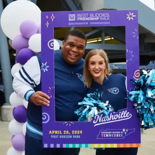 A man and woman with pom-poms pose in a handheld Best Buddies frame