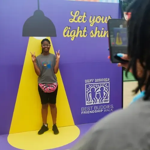 A girl poses with peace fingers in front of a sign with a spotlight graphic behind her that says
