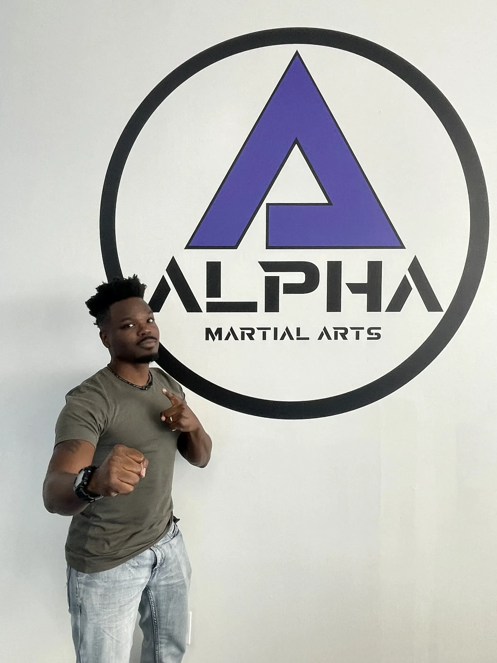 Alpha Martial Arts logo on the wall, with a person posing in front of it, pointing towards the camera