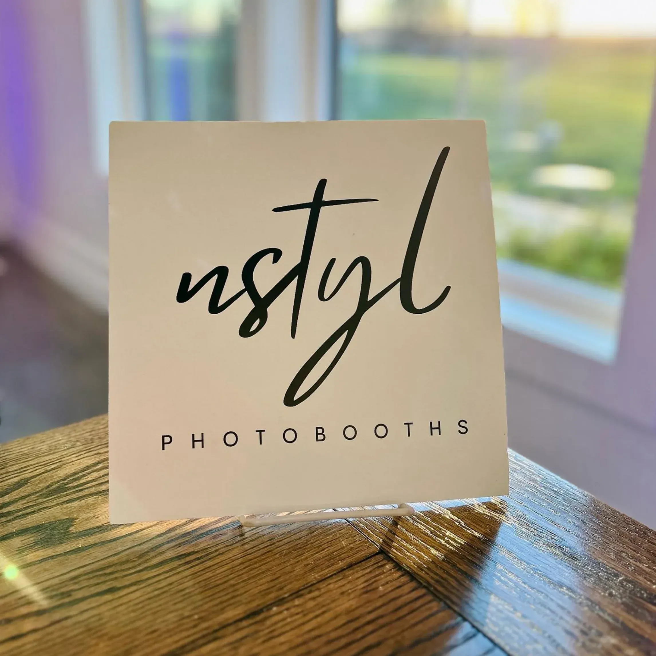 Small tabletop display sign with the Nstyl Photobooths logo in black script on a cream background, sitting on a wooden surface with soft daylight in the background.