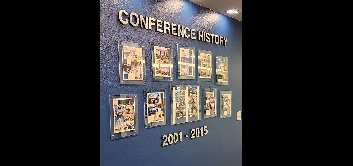 Wall-mounted conference history display featuring raised dimensional lettering and acrylic photo holders for a corporate interior space.