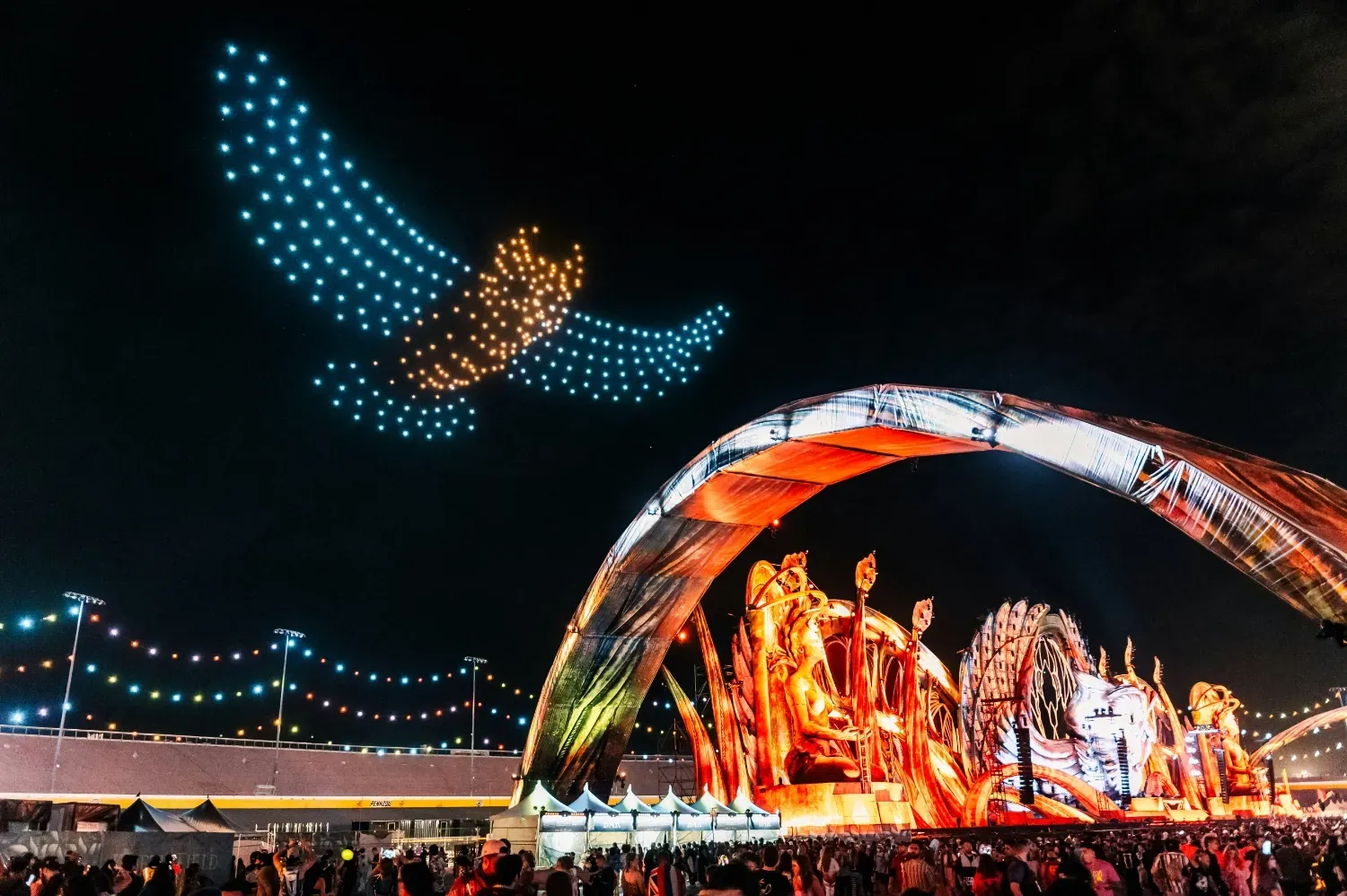 An owl made out of drones is flying in the sky above a festival.