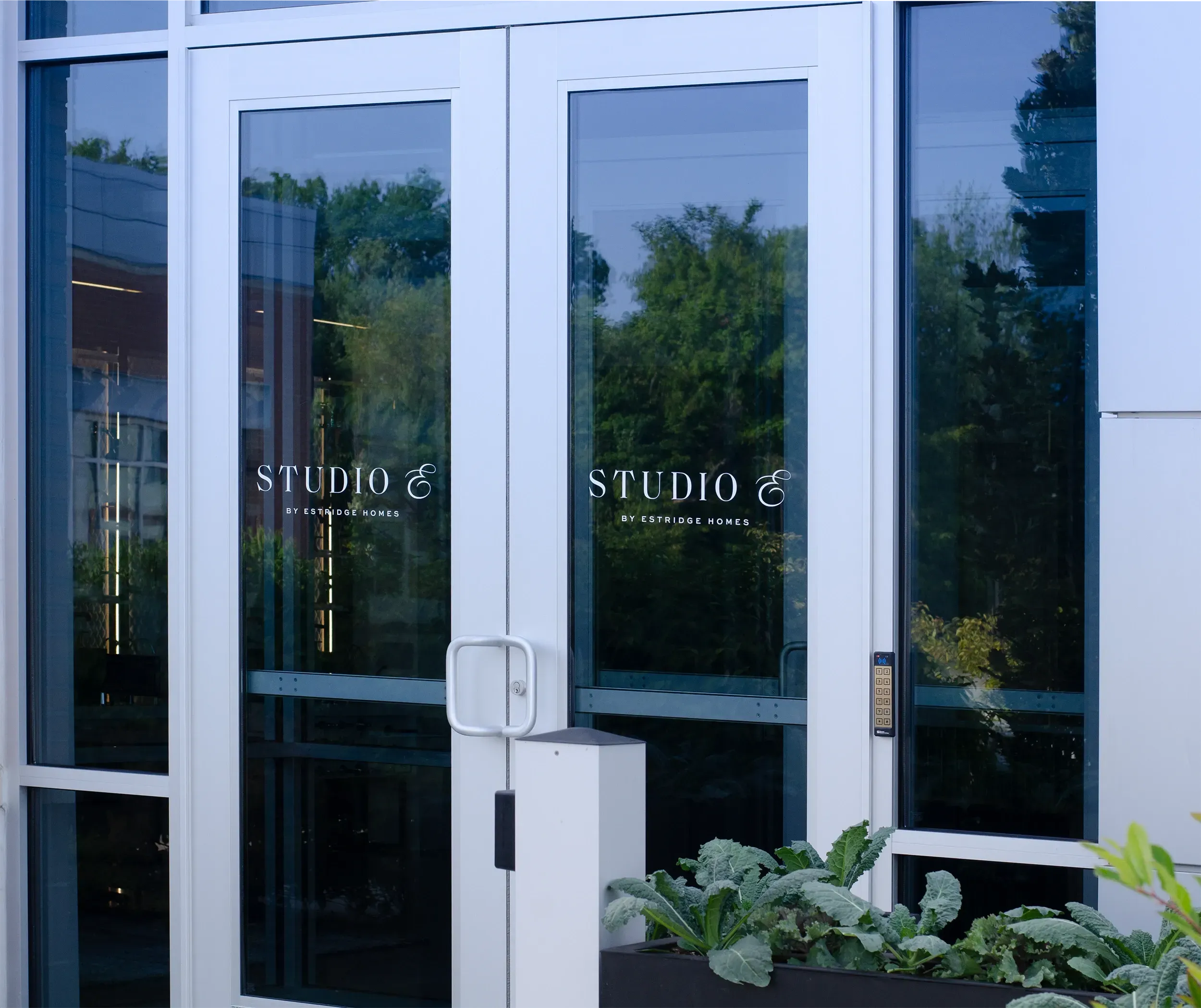 Entrance of Studio E by Estridge Homes featuring sleek glass doors with the Studio E logo in white lettering. The surrounding area includes greenery and a clean, minimalistic building design.