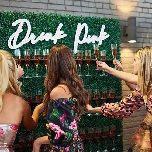 Women take champagne flutes from a greenery wall with a sign saying 