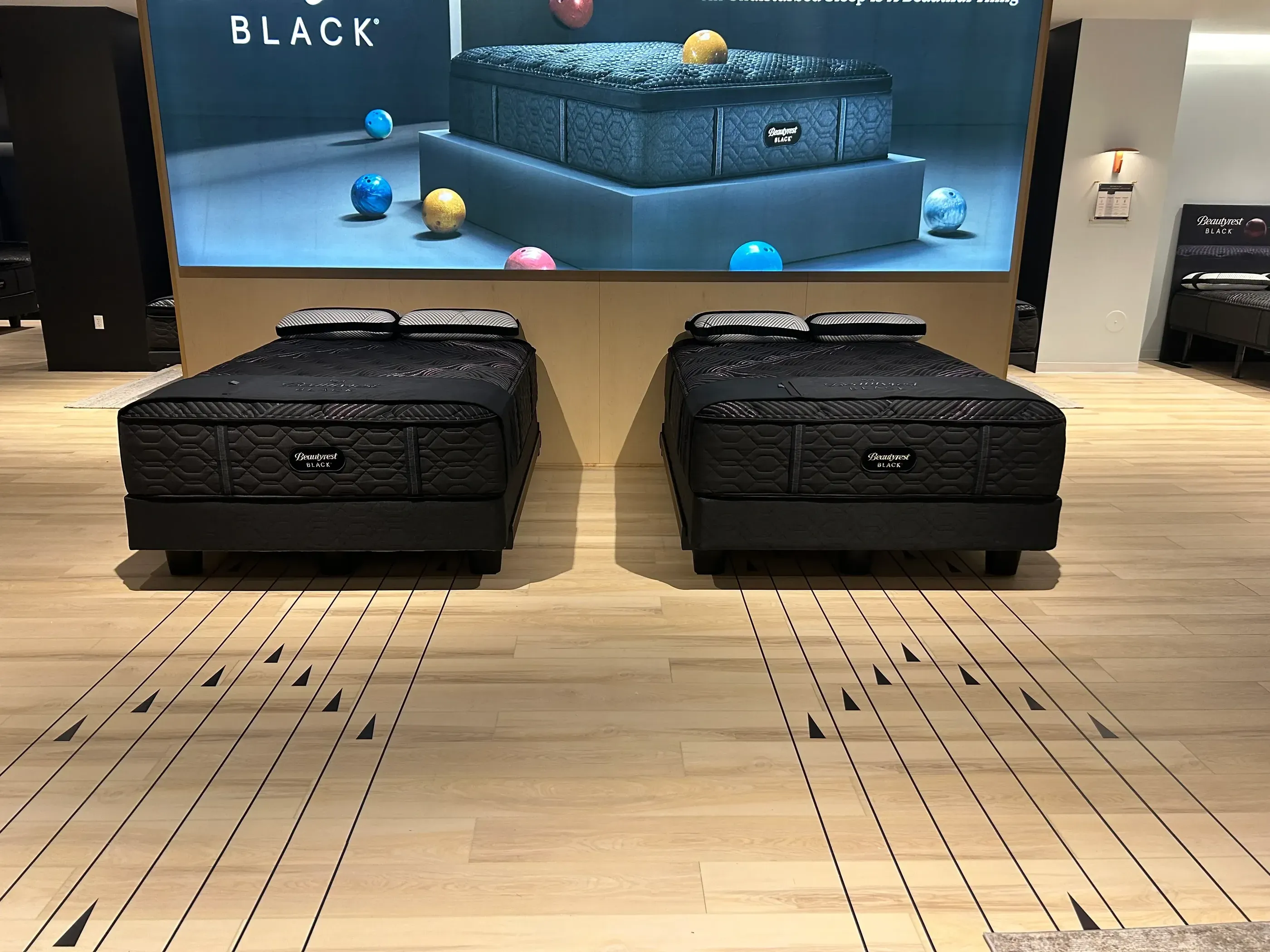Two Beautyrest Black mattresses displayed side by side on a light wooden floor, with branding and colorful spheres in the background.
