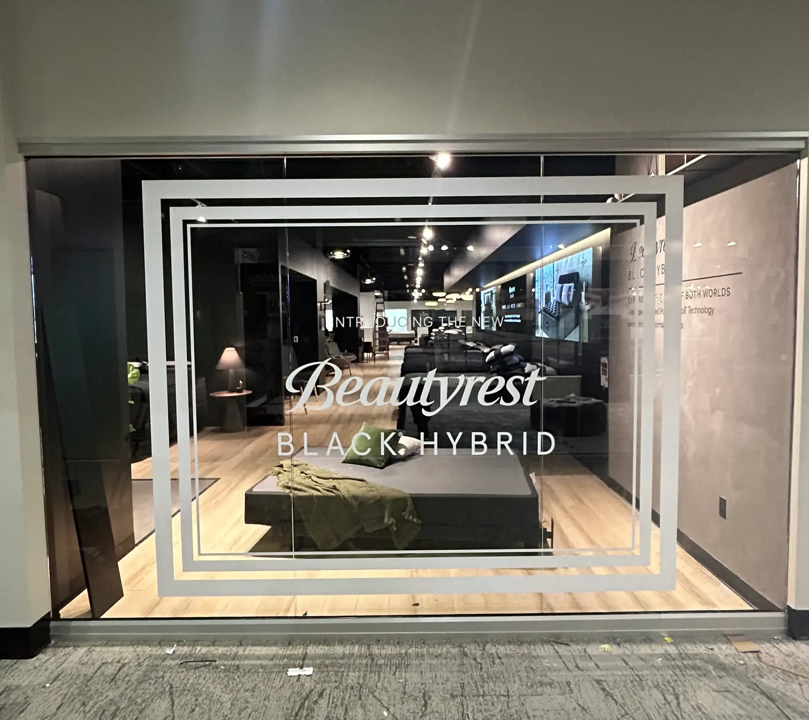 A storefront window featuring a Beautyrest Black Hybrid display, with white geometric designs framing the product branding.