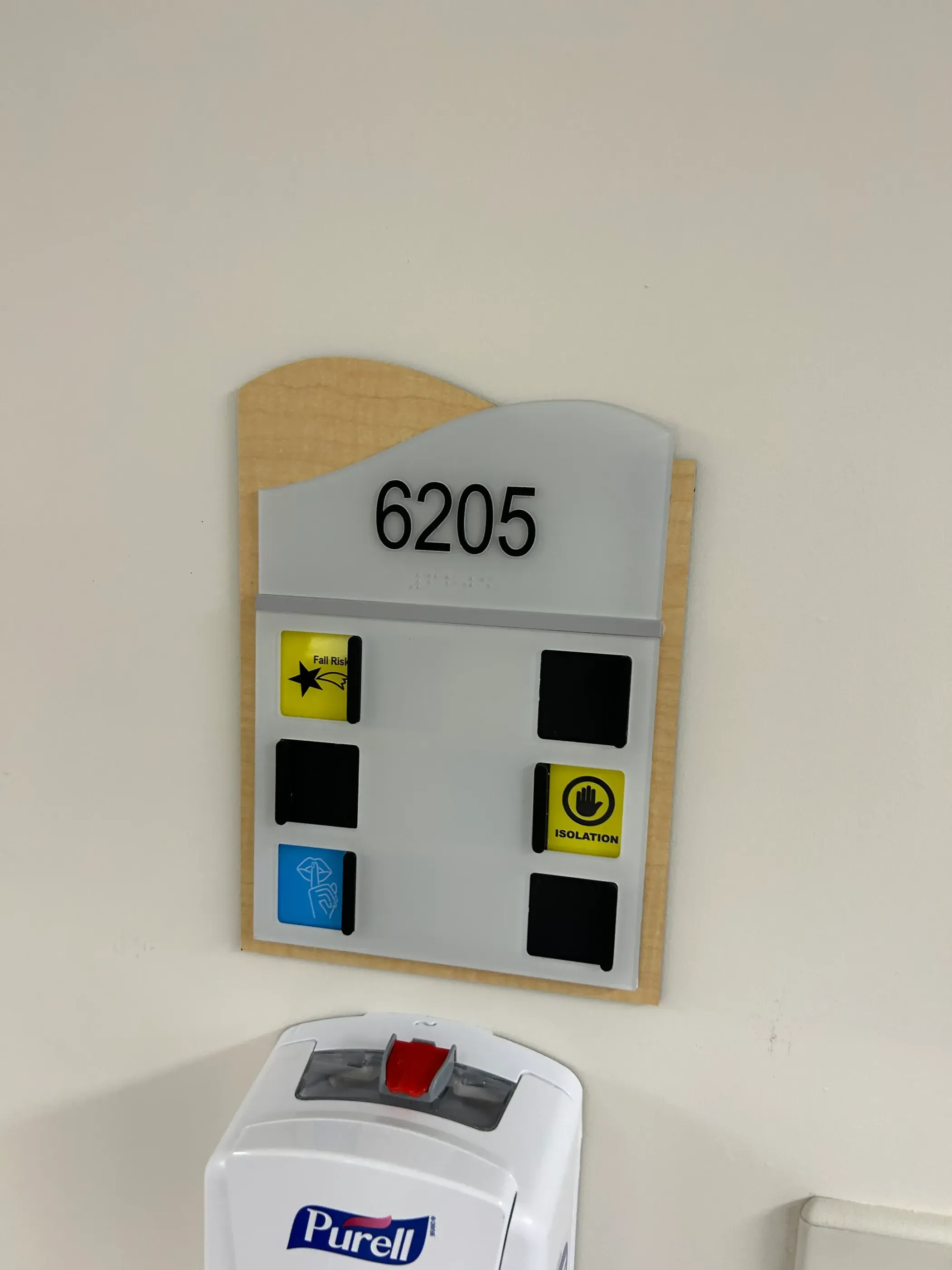 Room sign for hospital room 6205 at Sunrise Hospital, featuring a wooden plaque with a curved top. The sign includes slots for indicators such as ‘Fall Risk’ and ‘Isolation’ and Braille underneath the room number. A hand sanitizer dispenser is mounted below the sign.