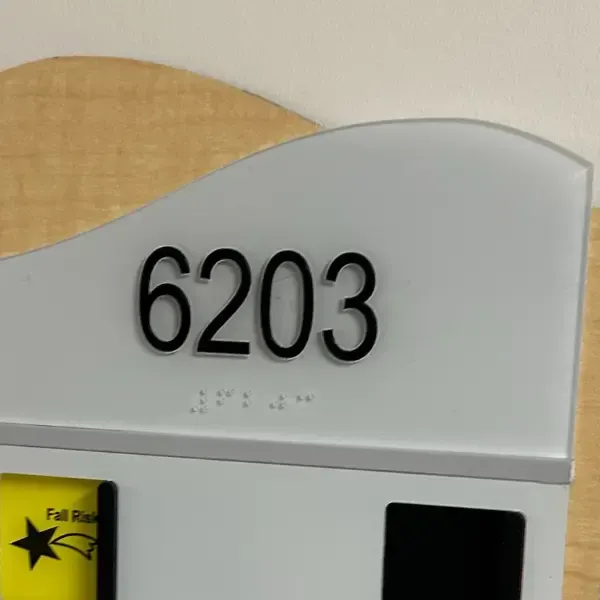 Close-up of the hospital room sign for room 6203 at Sunrise Hospital, featuring large black numbers and Braille beneath them. The sign includes a curved top and slots for status indicators like ‘Fall Risk’ on a wooden plaque.