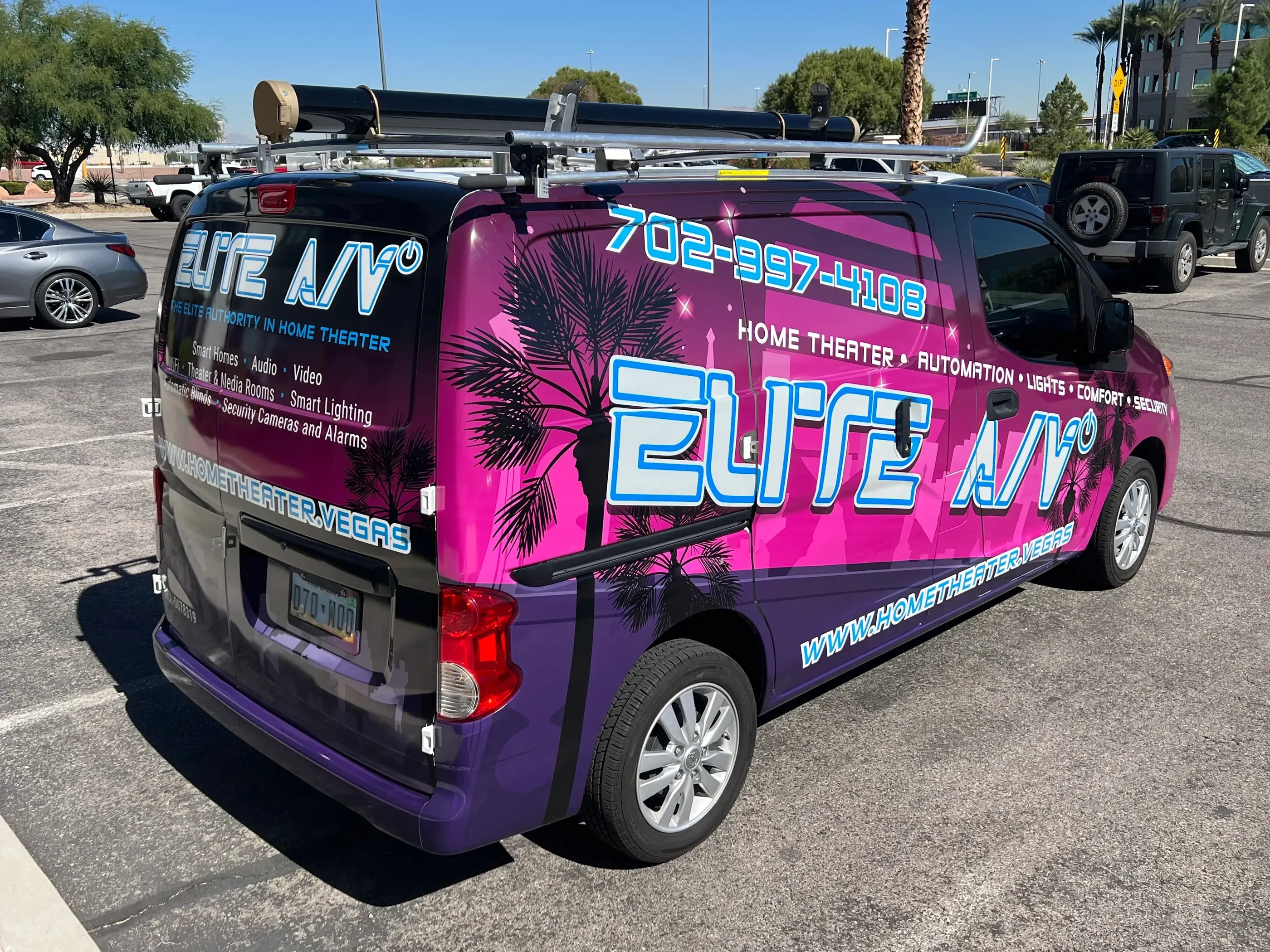 Purple van with Elite A/V branding on the side, detailing their services in home theater, lighting, and security. The design includes bright text and palm tree graphics, parked outside an AlphaGraphics location.